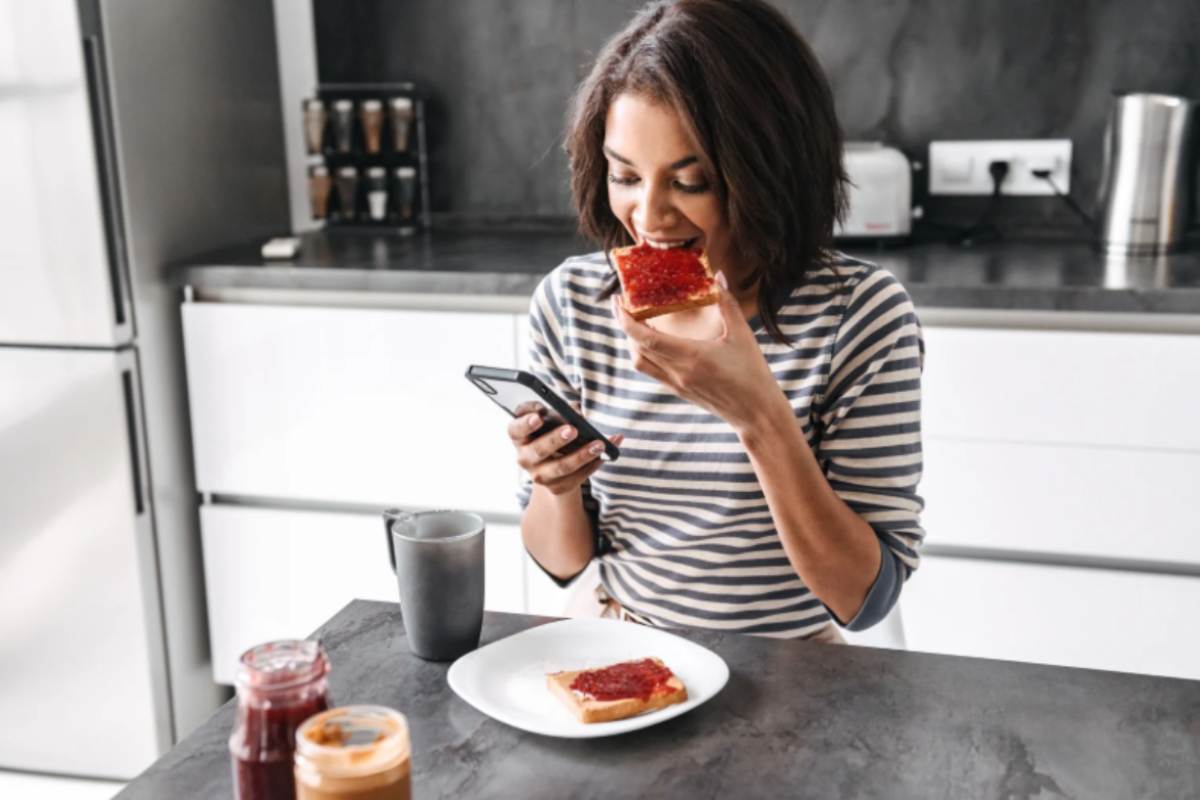 donna che mangia una fetta biscottata con marmellata mentre guarda il cellulare